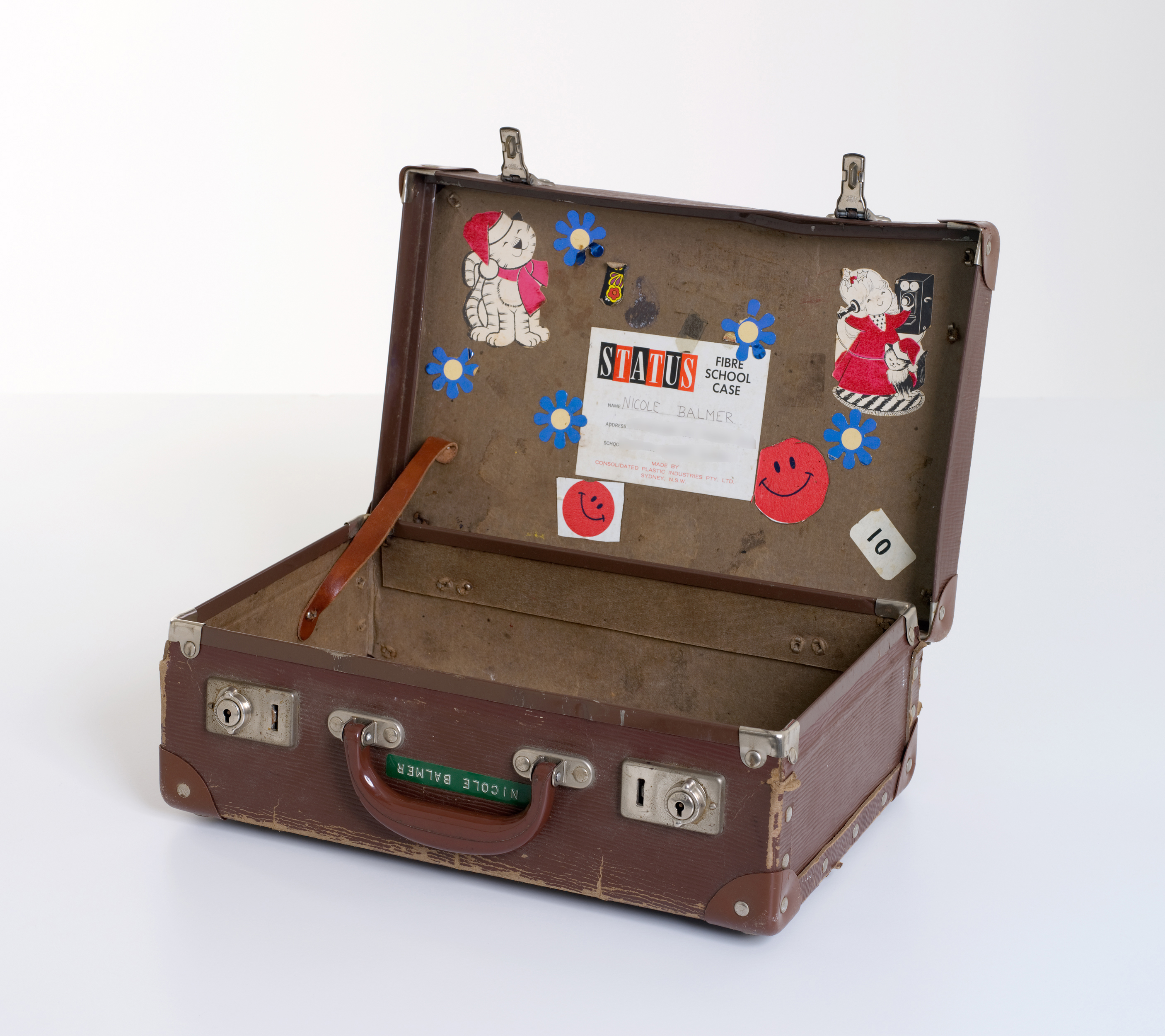 A brown, rectangular leather school case (open). The name ‘Nicole Balmer’ is marked next to the handle in Dymo tape, and the inside of the lid is covered in stickers.
